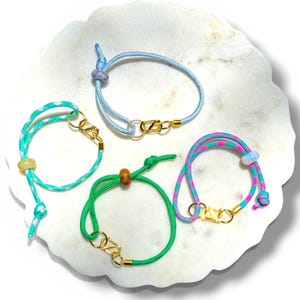 May include: Four colorful bracelets with gold-colored clasps and beads are displayed on a white, marble-like surface. The bracelets are in shades of blue, green, and pink, with varying cord patterns.
