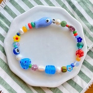 Pode incluir: Uma pulseira de contas multicolorida com uma variedade de formas e materiais, incluindo uma conta azul-clara em forma de peixe, uma conta azul em forma de coração com padrões florais brancos, uma conta de olho grego e contas em forma de flor.