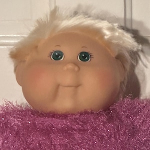 Può includere: Una bambola con viso rotondo, pelle chiara e guance rosate. Ha capelli biondi corti e lisci, occhi verdi con ciglia visibili e un naso piccolo. La bambola indossa un maglione viola brillante, testurizzato e soffice, con un leggero riflesso.