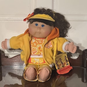 May include: A doll with dark brown hair and blue eyes wearing a yellow visor with an orange flower, a yellow hooded jacket, and a yellow floral print top and pants. The doll carries a small yellow bag featuring a graphic of hula dancers.