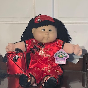 May include: A Cabbage Patch Kids doll with black yarn hair, wearing a red satin outfit with gold floral patterns and black trim. The doll has a matching red headband with a panda graphic and a red accessory bag featuring a panda and bamboo design.