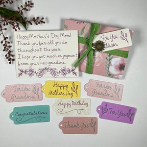 May include: A set of colourful gift tags with various messages, including "Happy Mother's Day", "Happy Birthday", "Thank You", and "For You". The tags are decorated with floral designs and have a glittery finish.