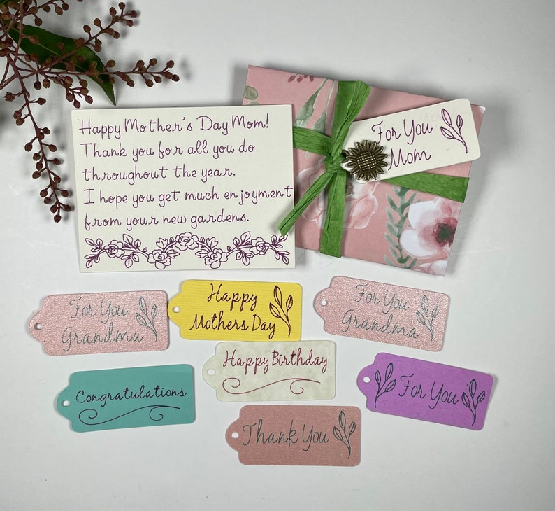 May include: A set of colourful gift tags with various messages, including "Happy Mother's Day", "Happy Birthday", "Thank You", and "For You". The tags are decorated with floral designs and have a glittery finish.