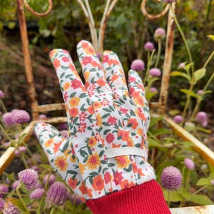 May include: A gardening glove with a white background, covered in a floral pattern of orange, yellow, and red flowers. The cuff is red, and a white elastic band secures the glove. The glove is set against a backdrop of a rusty metal chair and purple flowers.