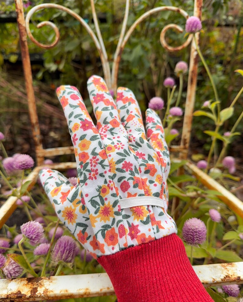 May include: A gardening glove with a white background, covered in a floral pattern of orange, yellow, and red flowers. The cuff is red, and a white elastic band secures the glove. The glove is set against a backdrop of a rusty metal chair and purple flowers.