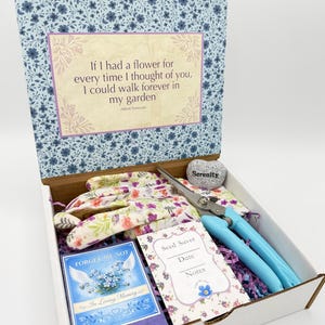 May include: A gardening gift box with a floral print lid and a quote. Inside are gardening gloves, blue handled shears, a seed saver, a 'Forget-Me-Not' card, and a stone with the word 'Serenity'.