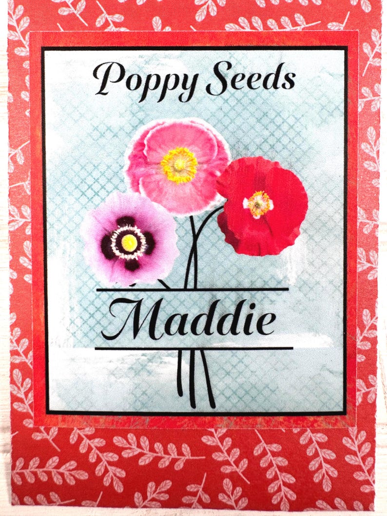 May include: A seed packet featuring three poppy flowers in shades of pink and red, with yellow centers, against a light blue background. The text "Poppy Seeds" and "Maddie" are displayed in elegant black script.