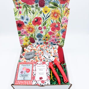 May include: A gardening gift box with a floral design on the lid and interior. The box contains gardening gloves, a rock with the word "Peace", red handled pruning shears, seed packets, and green paper filler.