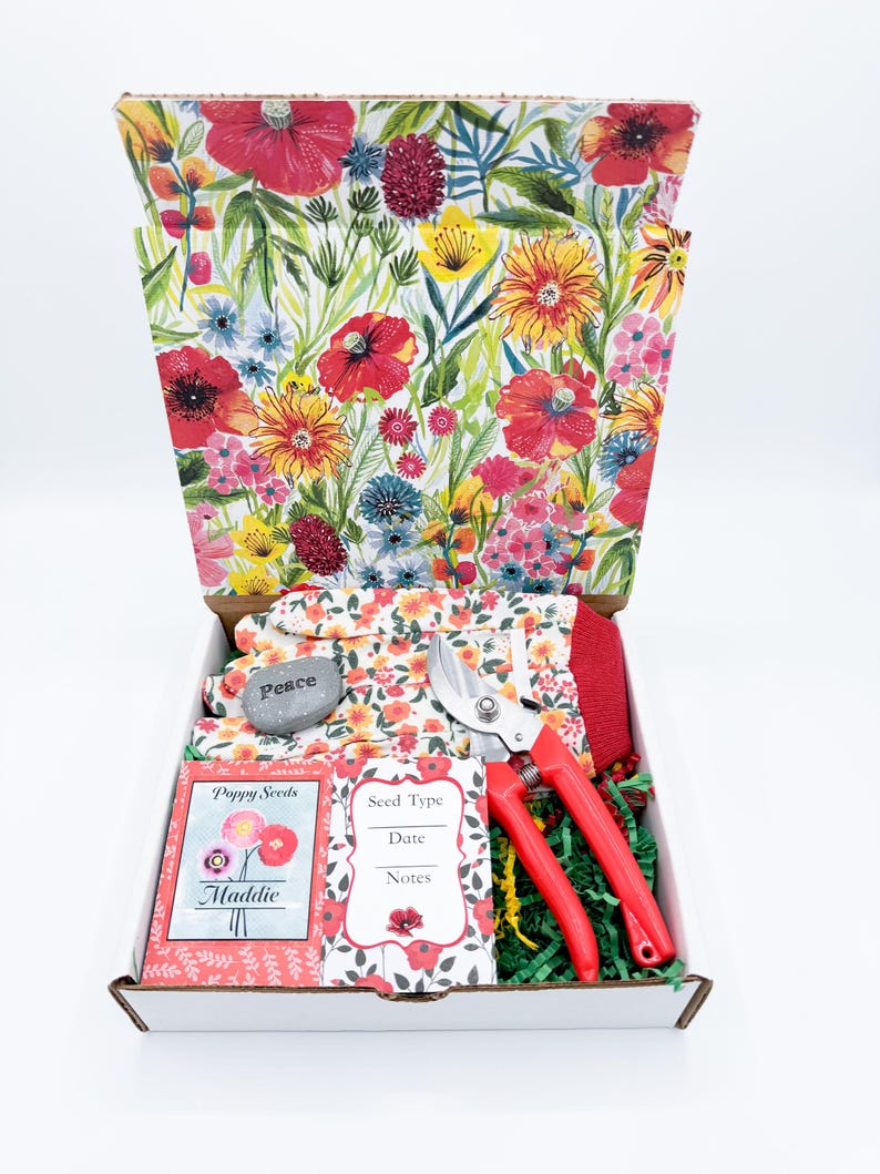 May include: A gardening gift box with a floral design on the lid and interior. The box contains gardening gloves, a rock with the word "Peace", red handled pruning shears, seed packets, and green paper filler.