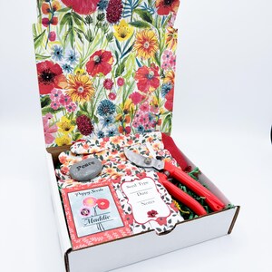 May include: A white box with a floral print lid, containing gardening tools and supplies. Includes red-handled pruning shears, gardening gloves, seed packets, and a stone with the word "Peace". The box is filled with green packing material.