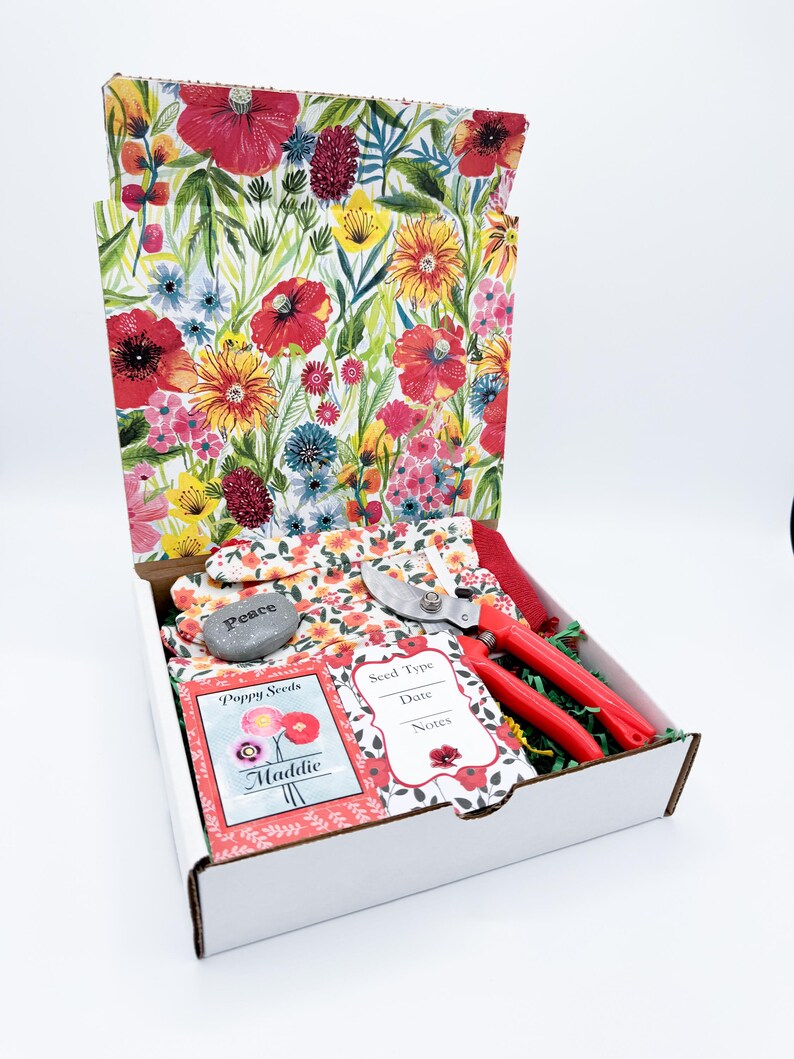 May include: A white box with a floral print lid, containing gardening tools and supplies. Includes red-handled pruning shears, gardening gloves, seed packets, and a stone with the word "Peace". The box is filled with green packing material.