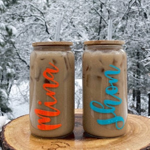May include: Two clear glass mason jars with bamboo lids and straws. The jars are filled with iced coffee and have personalized names, "Mina" in orange and "Gwen" in blue, written on the sides.