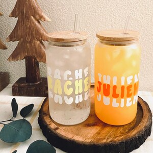 May include: Two glass mason jars with bamboo lids and straws. The jars are filled with beverages. One jar has a yellow and white design that says "Rache" and the other jar has an orange and white design that says "Julien".