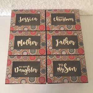 K&ouml;nnte beinhalten: Sechs kleine Geschenkboxen mit einem braunen und wei&szlig;en Schneeflockenmuster. Die Boxen haben ein transparentes Fenster auf der Vorderseite und jede Box tr&auml;gt eine andere Inschrift. Die Inschriften sind: "Jessica", "Frohe Weihnachten", "Mutter", "Vater", "An Meine Tochter", und "An Meinen Sohn".