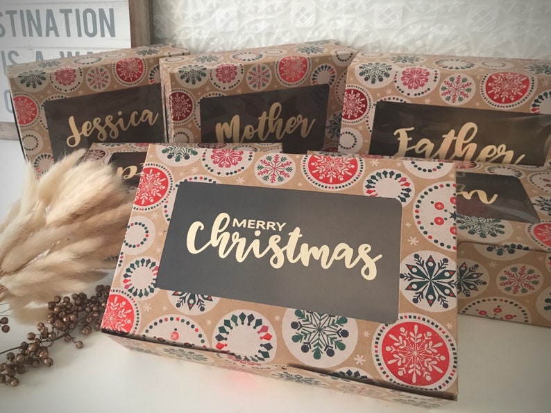 K&ouml;nnte beinhalten: Vier braune Geschenkboxen mit einem Schneeflockenmuster und durchsichtigen Fenstern. Die Boxen sind mit "Jessica", "Mutter", "Vater" und "Frohe Weihnachten" beschriftet.