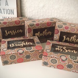 K&ouml;nnte beinhalten: F&uuml;nf kleine Geschenkboxen mit einem Schneeflockenmuster. Die Boxen haben transparente Fenster, die personalisierte Botschaften zeigen, darunter "Jessica", "Mutter", "Vater", "An meine Tochter" und "An meinen Sohn".