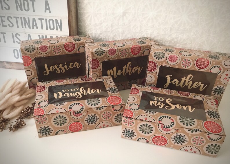 K&ouml;nnte beinhalten: F&uuml;nf kleine Geschenkboxen mit einem Schneeflockenmuster. Die Boxen haben transparente Fenster, die personalisierte Botschaften zeigen, darunter "Jessica", "Mutter", "Vater", "An meine Tochter" und "An meinen Sohn".