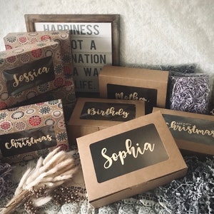 K&ouml;nnte beinhalten: Eine Sammlung brauner Karton-Geschenkboxen mit durchsichtigen Fenstern. Die Boxen sind mit verschiedenen Mustern verziert und haben goldene Beschriftungen. Die Beschriftungen beinhalten Namen wie "Jessica", "Sophia", "Geburtstag" und "Weihnachten".