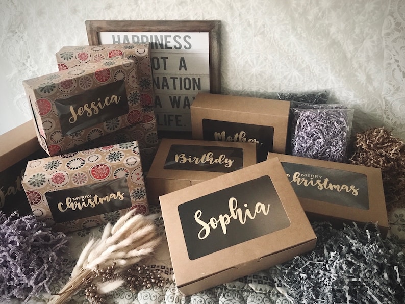 K&ouml;nnte beinhalten: Eine Sammlung brauner Karton-Geschenkboxen mit durchsichtigen Fenstern. Die Boxen sind mit verschiedenen Mustern verziert und haben goldene Beschriftungen. Die Beschriftungen beinhalten Namen wie "Jessica", "Sophia", "Geburtstag" und "Weihnachten".