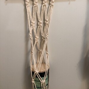 May include: A white macrame plant hanger with a wooden ring at the top and a glass jar with a black label that says "Stress Relief" hanging from it.