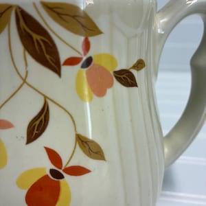 May include: Cream-colored ceramic pitcher with a gold rim and a curved handle. The pitcher is decorated with a floral pattern in shades of yellow, orange, and brown. The pitcher has vertical ridges and a smooth, glossy finish.