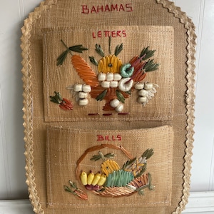 May include: A woven wall organizer with two pockets, labeled "Letters" and "Bills". The pockets are decorated with colorful straw flowers and fruit.