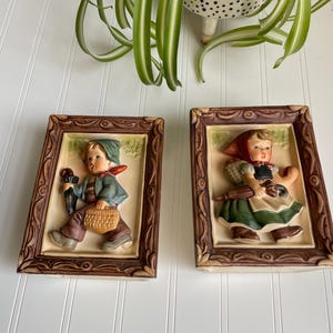 May include: A pair of vintage framed wall plaques featuring 3D figurines of a boy and a girl in traditional German clothing. The boy is holding an umbrella and a basket, while the girl is holding a basket and a rake. The plaques have a brown wooden frame with a decorative design.