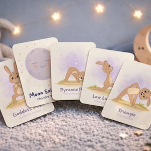 May include: A set of five yoga cards featuring illustrations of a cartoon bunny demonstrating yoga poses. The cards include the poses: Goddess Pose, Moon Salutation, Pyramid Pose, Low Lunge, and Triangle. The background includes a plush bunny, a crescent moon, and star-shaped lights.