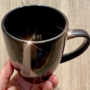 Puede incluir: Una taza de cer&aacute;mica negra brillante con un asa curva. La taza tiene una superficie lisa y reflectante. La taza est&aacute; sostenida por una mano, y el fondo es una veta de madera clara. La taza parece estar vac&iacute;a.