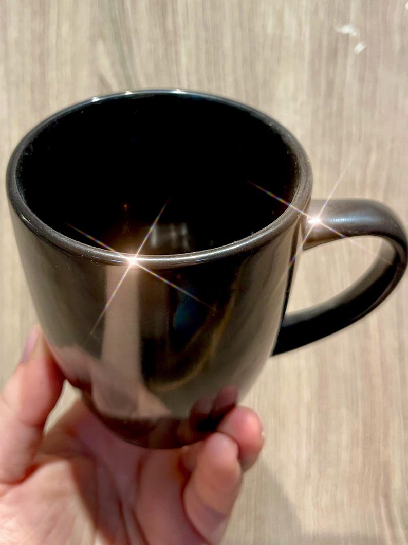 Puede incluir: Una taza de cer&aacute;mica negra brillante con un asa curva. La taza tiene una superficie lisa y reflectante. La taza est&aacute; sostenida por una mano, y el fondo es una veta de madera clara. La taza parece estar vac&iacute;a.