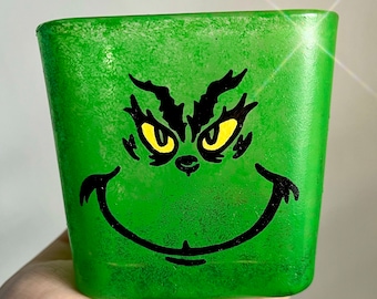 May include: Green ceramic planter with a black and yellow Grinch face painted on the front. The Grinch is smiling with his eyes wide open.