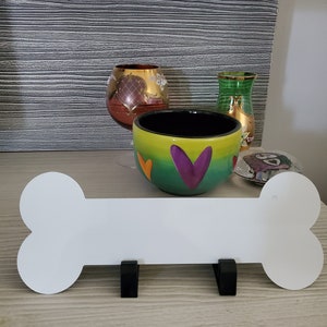 May include: White ceramic bone-shaped sign with two black metal hooks. The sign is sitting on a wooden surface. There are two decorative vases and a bowl with colorful hearts on the surface behind the sign.