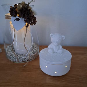 Könnte beinhalten: Eine weiße Keramikbox mit einer Teddybär-Figur. Der Deckel hat ein dekoratives Muster, und die Seiten haben kleine, beleuchtete Löcher. Ein Glas mit einer Kerze und getrockneten Blumen ist ebenfalls zu sehen.