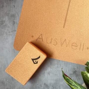 May include: A cork yoga block with a black logo on it. The block is sitting on a cork yoga mat with the text "Auswell" printed on it.
