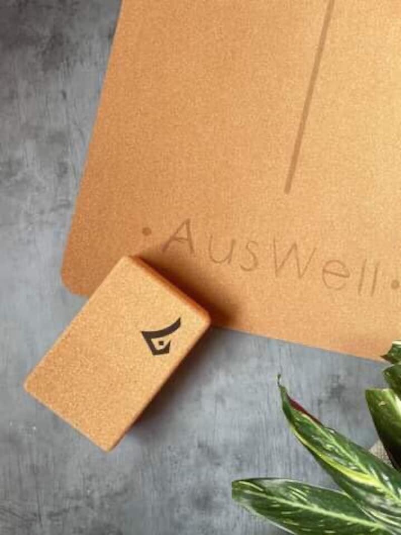 May include: A cork yoga block with a black logo on it. The block is sitting on a cork yoga mat with the text "Auswell" printed on it.