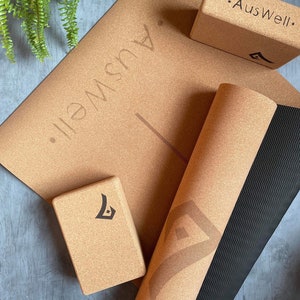 May include: A set of cork yoga accessories including a yoga mat, a yoga block, and a yoga bolster. The yoga mat has a black and brown striped pattern on the underside. The yoga block and bolster have the text "AusWell." printed on them.
