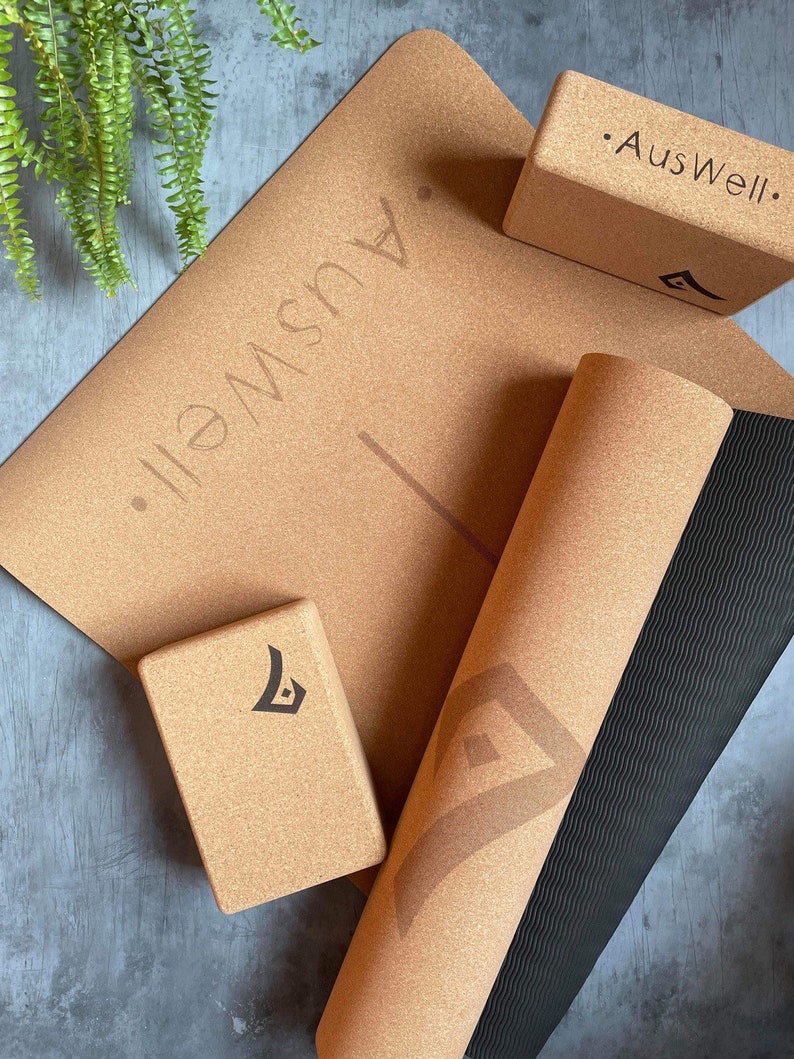 May include: A set of cork yoga accessories including a yoga mat, a yoga block, and a yoga bolster. The yoga mat has a black and brown striped pattern on the underside. The yoga block and bolster have the text "AusWell." printed on them.