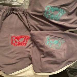 May include: Two pairs of gray athletic shorts with white trim. One pair has a red glitter "BO$$ GIRL" logo and the other pair has a teal glitter "BO$$ GIRL" logo.
