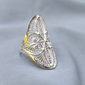 May include: A silver filigree ring with a delicate floral design. The ring is oval shaped and has a wide band.