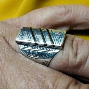 May include: A silver ring with a rectangular design featuring intricate geometric patterns and black lines. The ring is worn on a finger.