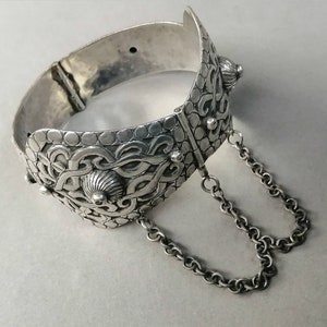 May include: A silver bracelet with intricate knotwork designs and two dangling chains. The bracelet is hinged and has a textured surface.