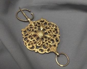 May include: A gold-toned metal brooch with an intricate, ornate design. The brooch features a central oval-shaped piece and a decorative loop at the top.