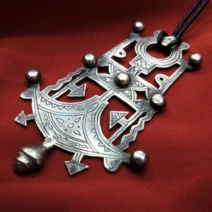 May include: A silver pendant with intricate geometric designs and a black cord. The pendant is shaped like a cross with a circle at the top and a pointed bottom.