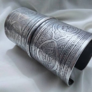 May include: Three silver bracelets with intricate embossed designs. The bracelets are stacked on top of each other and feature a repeating pattern of stylized fish and geometric shapes.