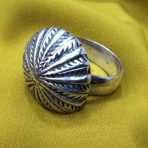May include: A silver ring with an oval, textured design. The ring's band is smooth and simple, while the oval top features a detailed, radiating pattern. The ring is set against a yellow background.