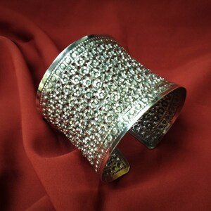 May include: A silver cuff bracelet with a delicate floral design. The bracelet is made of intricate metalwork and has a wide, open design.