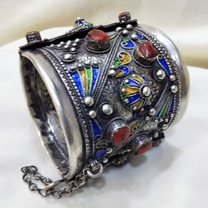 May include: A silver bracelet with intricate enamel work in blue, green, and yellow. The bracelet is decorated with red coral stones and has a chain attached.