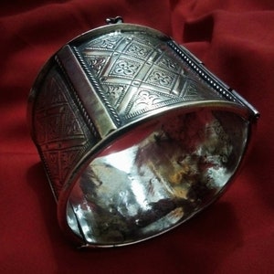 May include: A silver bracelet with intricate geometric designs and a hinged clasp. The bracelet is made of metal and has a polished finish.