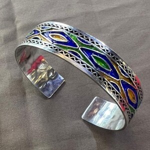 May include: A silver bangle bracelet with a geometric pattern in blue, green, and gold enamel.