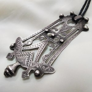 May include: A silver pendant with intricate geometric designs, featuring a central open space and a loop at the top for attaching a cord or chain. The pendant is made of silver and has a dark patina.